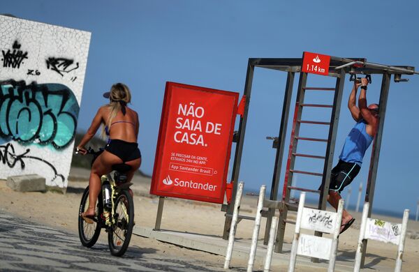 Девушка проезжает мимо баннера Не выходи из дома на пляже Ипанема в Рио-де-Жанейро во время пандемии коронавируса 