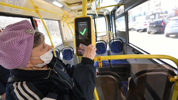 Женщина в защитной маске прикладывает социальную карту москвича к валидатору в салоне городского автобуса в Москве