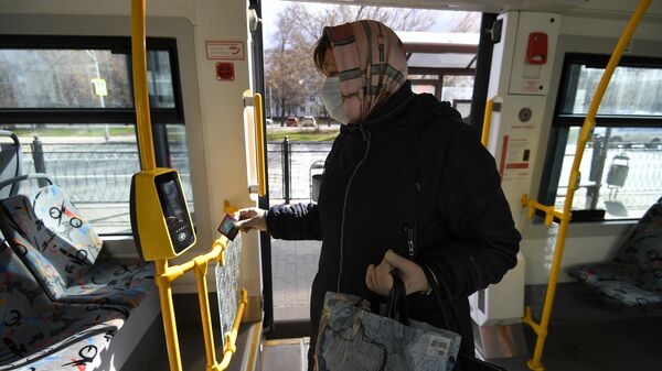 Женщина в защитной маске прикладывает социальную карту москвича к валидатору в салоне городского автобуса в Москве