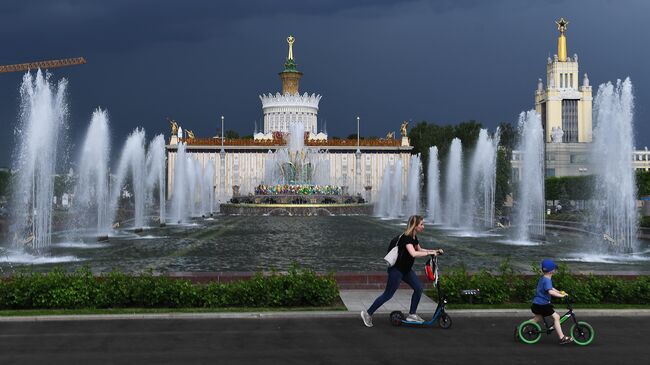 Посетители ВДНХ катаются на самокатах у фонтанов. В центре: фонтан Каменный цветок и павильон № 58