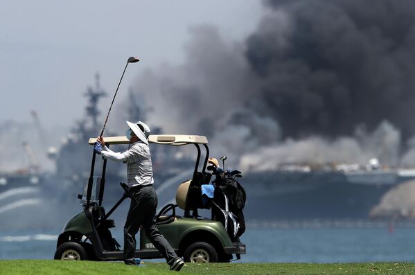 Мужчина играет в гольф на фоне горящего универсального десантного корабля USS Bonhomme Richard (LHD 6)