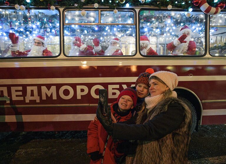 Прохожие фотографируются на фоне праздничного Дедморобуса с музыкантами в костюмах Дедов Морозов, который ездит по улицам Санкт-Петербурга в преддверии нового года