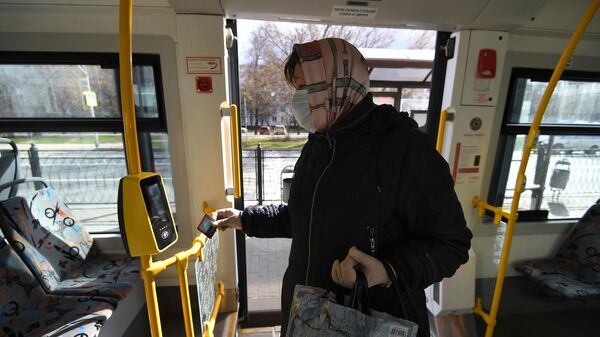 Женщина в защитной маске прикладывает карту к валидатору в салоне автобуса