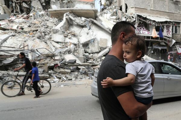 Палестинец с ребенком проходит мимо разрушенных в результате бомбардировки зданий в секторе Газа, после объявления о прекращении огня между Израилем и ХАМАС