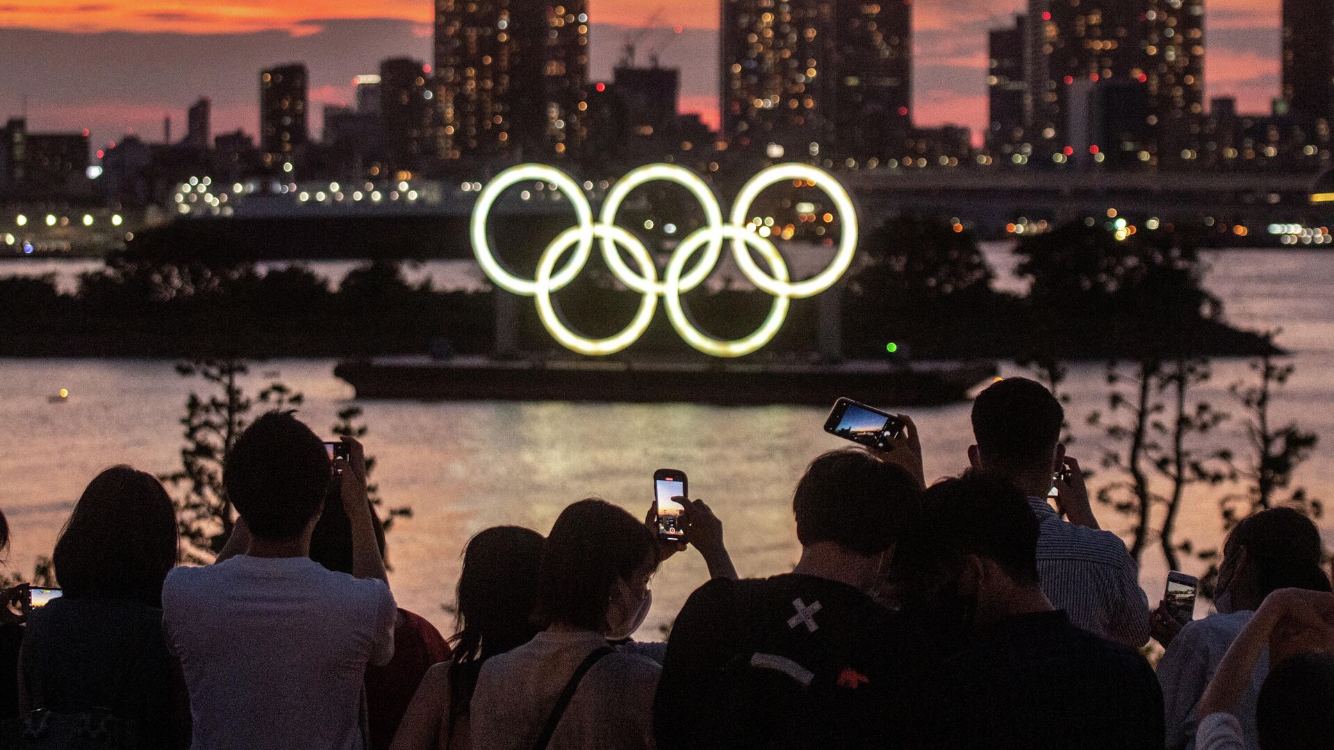 People take pictures as the Olympic rings lit up at dusk on the Odaiba waterfront in Tokyo on July 22, 2021 on the eve of the start of the Tokyo 2020 Olympic Games. (Photo by Philip FONG / AFP) - РИА Новости, 1920, 22.07.2021