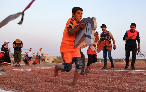 Перемещенные сирийские дети бегают с деревянными лошадиными головами во время Олимпиады 2020 года в лагере в городе Фуаа, на северо-западе Сирии