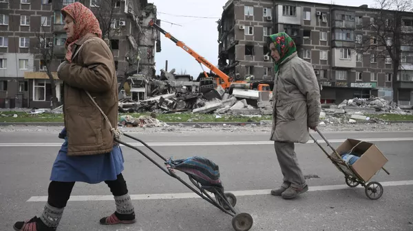 Mariupol lakói a város utcáin sétálnak.  A háttérben: a DPR Rendkívüli Helyzetek Minisztériumának munkatársai takarítják el a romokat a lerombolt házak közelében