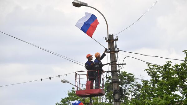 Сотрудники предприятия электрических сетей наружного освещения Горсвет вешают российский флаг на фонарный столб в Мариуполе
