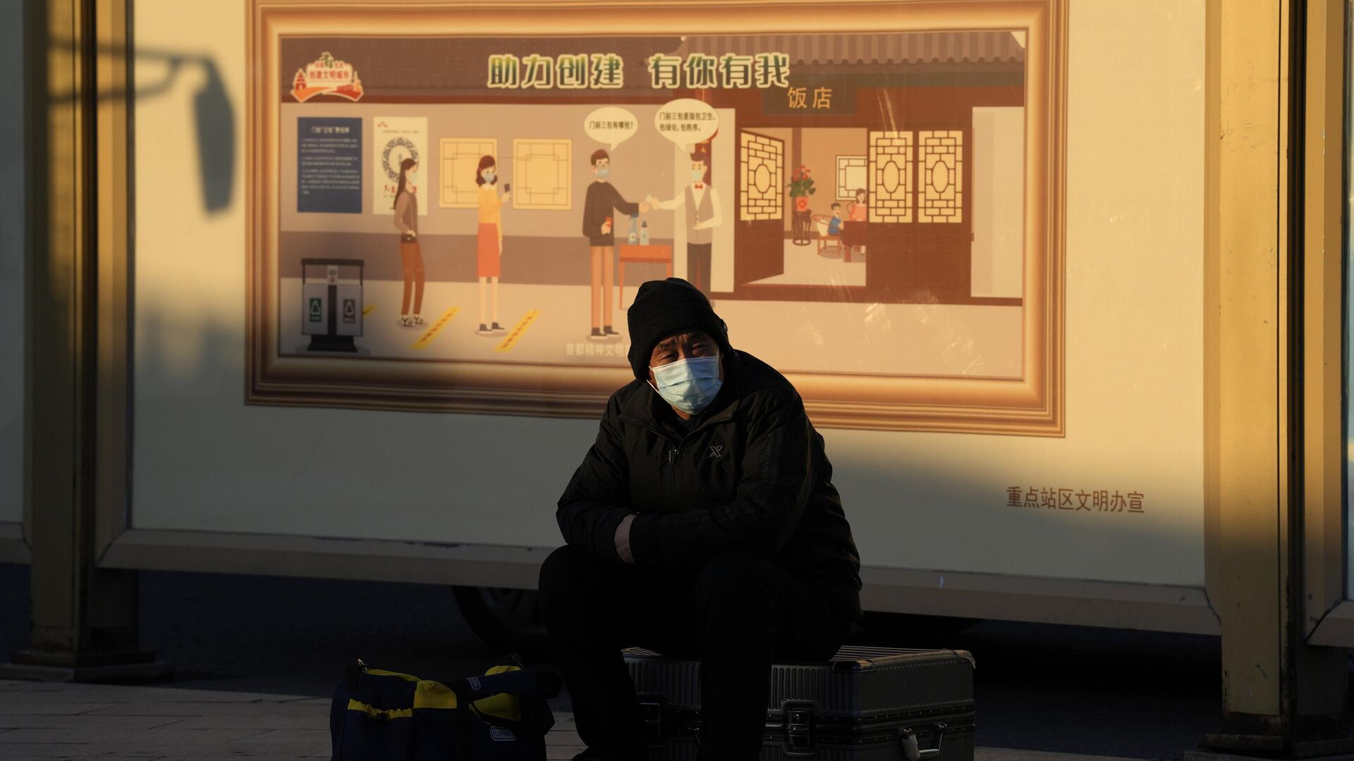 Мужчина в защитной маске сидит на своем багаже рядом с плакатом на тему COVID-19 в Пекине - РИА Новости, 1920, 08.12.2022