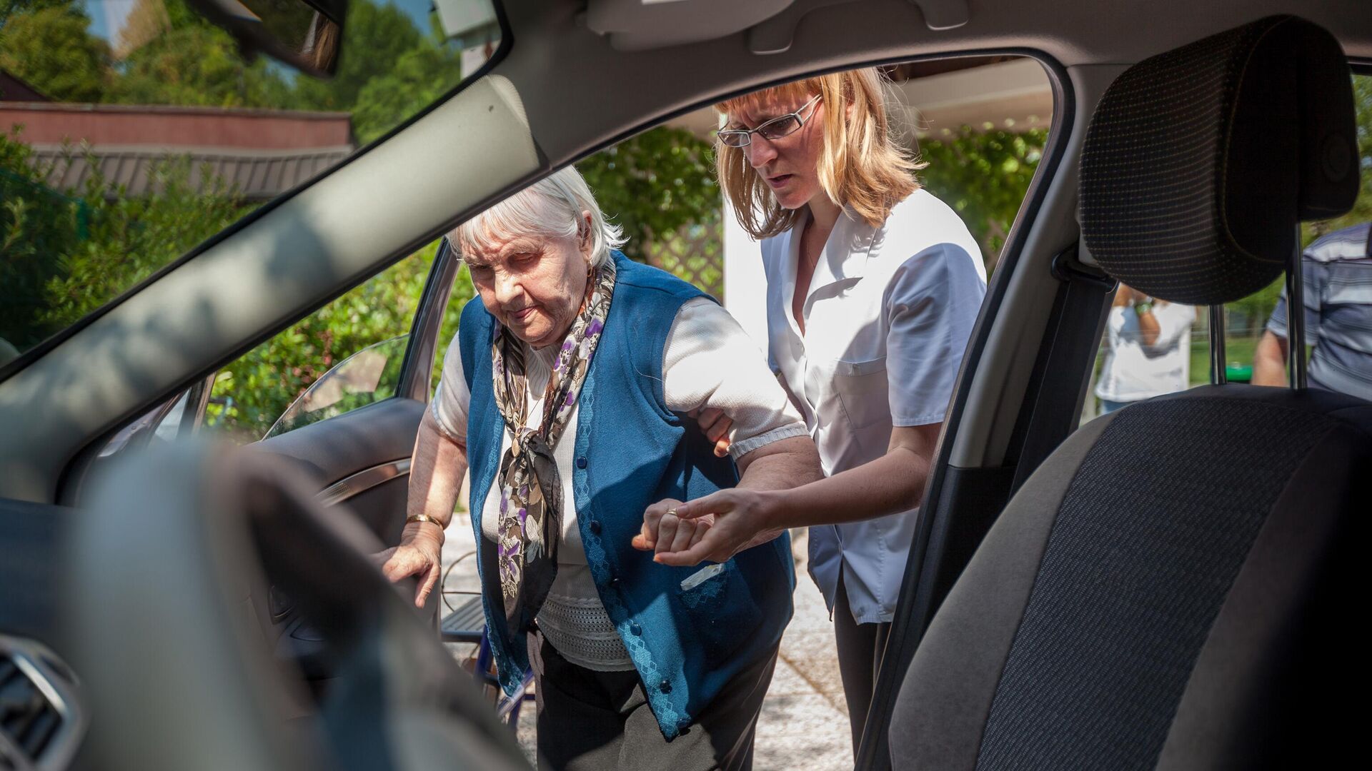 Медработник помогает пожилой женщине сесть в машину - РИА Новости, 1920, 11.07.2023