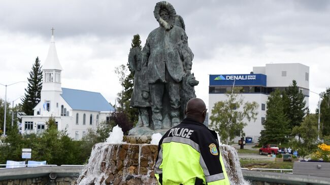 Сотрудник полиции у памятника первым поселенцам Аляски на набережной Чены и площади Голден Харт в городе Фэрбанкс на Аляске