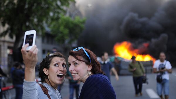 Туристы фотографируются на фоне горящего контейнера во время майской демонстрации в Барселоне, Испания
