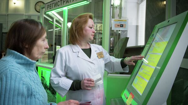 Сотрудница поликлиники помогает пациентке оформить талон на прием к врачу в терминале электронной очереди