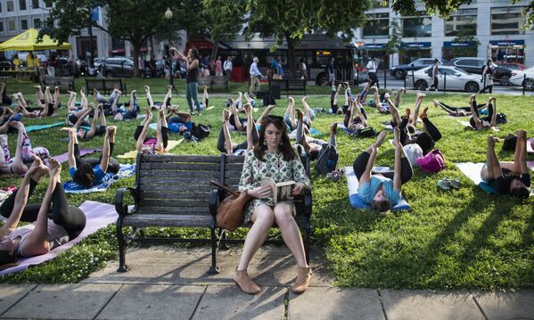 Женщина читает книгу на фоне группы, практикующей йогу в парке. Вашингтон, США