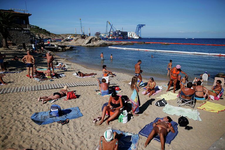 Люди отдыхают на пляже рядом с круизным лайнером Costa Concordia, где идет операция по его подъему